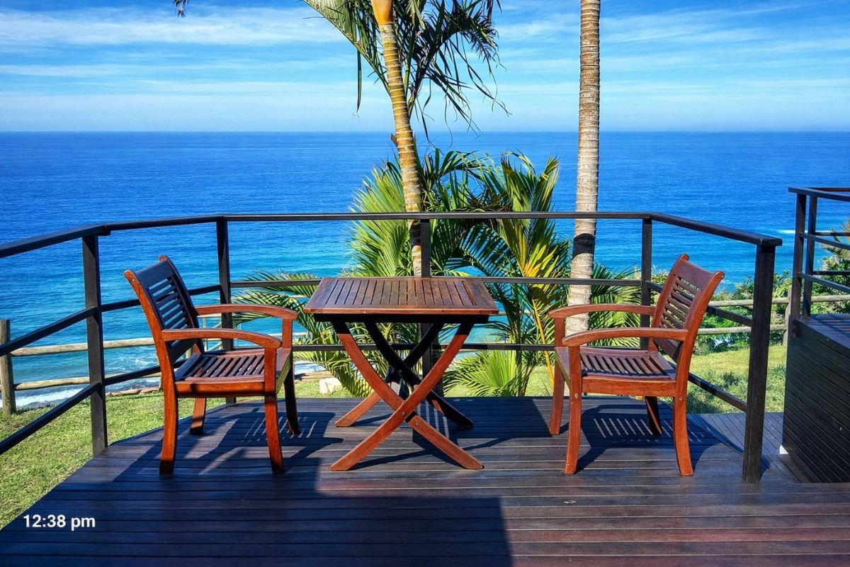 Enjoy a Bluff sea-view deck with wooden chairs and table, palm trees, and blue Indian Ocean horizon framed by railings at The Tides Inn B&amp;B.